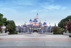 Sleeping Beauty's Castle with Blue Roof, pink on the outside of the main castle, gray stone walls, blue sky with light cloud cover, lights on black poles, blue, light blue and gold banners, trees with green leaves, pink flowers on a few trees,