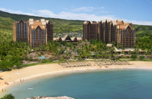 Disney's Aulani Resort with two tall towers, a main building that is 1 story with a lagoon and blue water, sand on the beach with chairs, along with different pools, buildings and palm trees on the property, blue skies with mountain greenery and light clouds