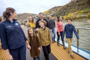 Family on a river cruise in Europe with fall colors of the trees and grasses in the background, all dressed in winter or fall like clothing, two sets of parents and four kids, and an adventures by disney cast member