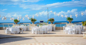 Wedding Reception Shown outdoors with a stone wall with four planters with flowers, star lights hanging from a pole, six tables with white tablecloths, white chairs and dinner settings for the guests