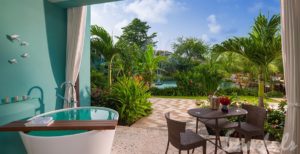 Lovers Lagoon Hideaway Walkout Jr Suite with Soaking Tub at a Sandals Resort, with a small table to dine at, curtains on patio, blue pool in the background, lush amount of green trees and bushes with a blue sky