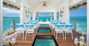 A Wedding Chapel over the water at a Sandals location in the caribbean with the blue waters in the background, white chairs with light blue cloth on the back of each chair, glass squares with the water below, a table and two pilars with beautiful floral arrangements