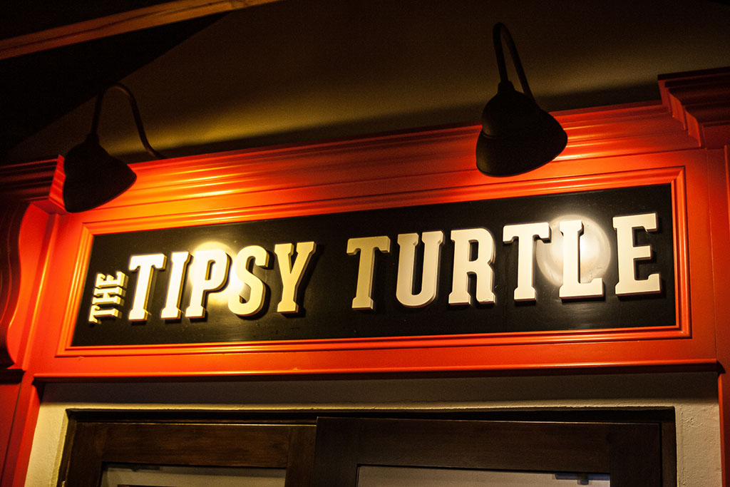 Outdoor sign at night with red border that says in white letters The Tipsy Turtle and two lights shining on sign